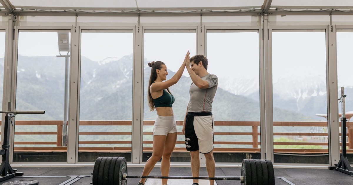 A fit couple celebrates a weightlifting success with a high-five in a bright, modern gym overlooking mountains.