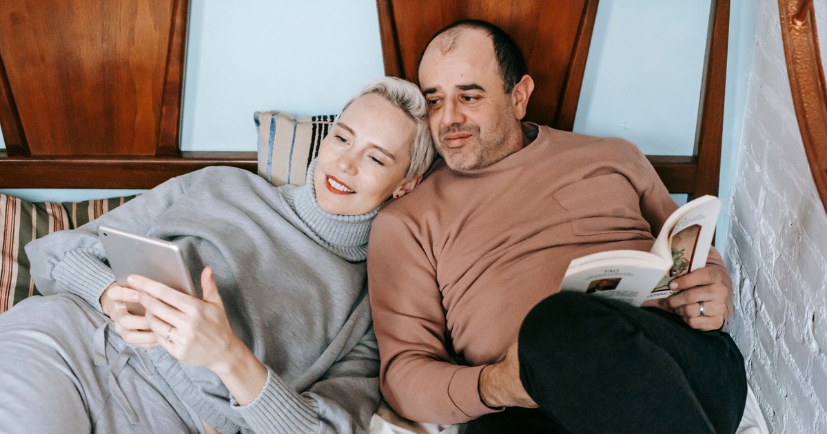 High angle of happy adult woman using tablet and cuddling with ethnic husband reading interesting book on bed at home