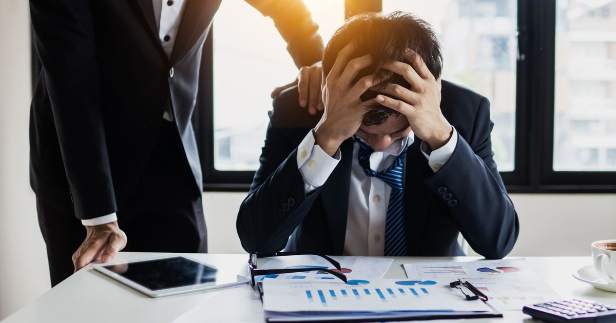 An overwhelmed businessman grasps his head, surrounded by reports in a bright office.