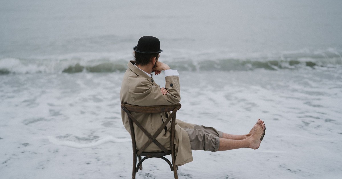 Man in coat and hat sitting on a chair at the edge of the ocean, lost in thought.