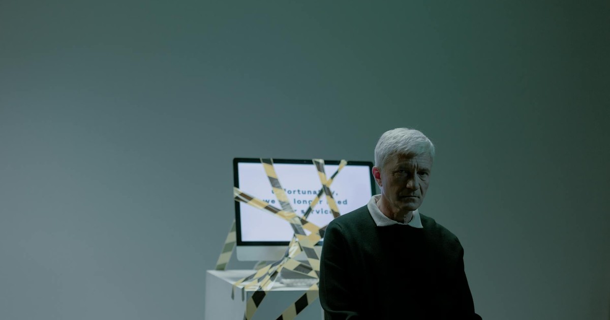 A senior adult sits indoors next to a computer wrapped in caution tape, symbolizing job loss and unemployment.