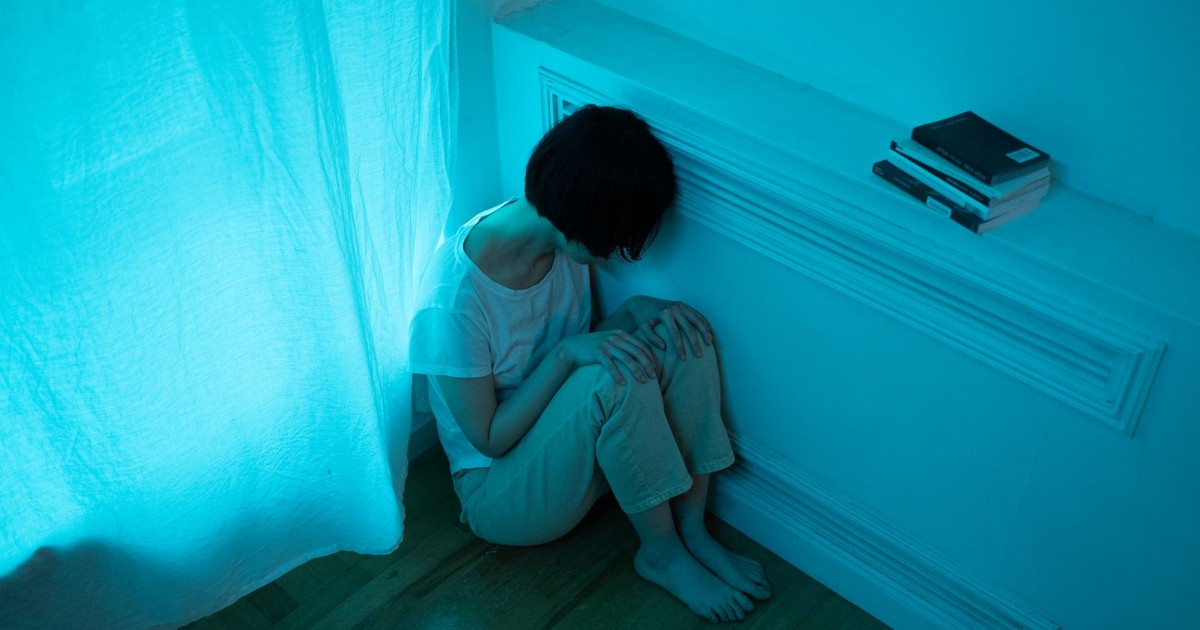 A woman in pajamas sits alone in a dimly lit room, expressing emotions of solitude.