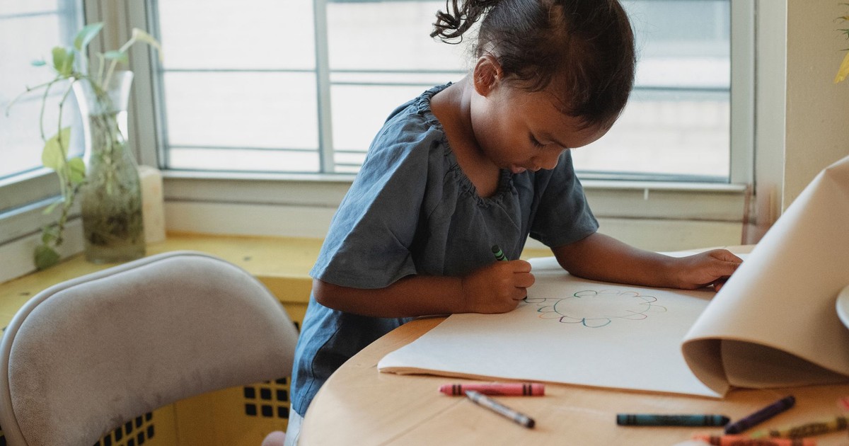 Concentrated Hispanic girl in casual clothes sitting at table and drawing picture with wax crayons