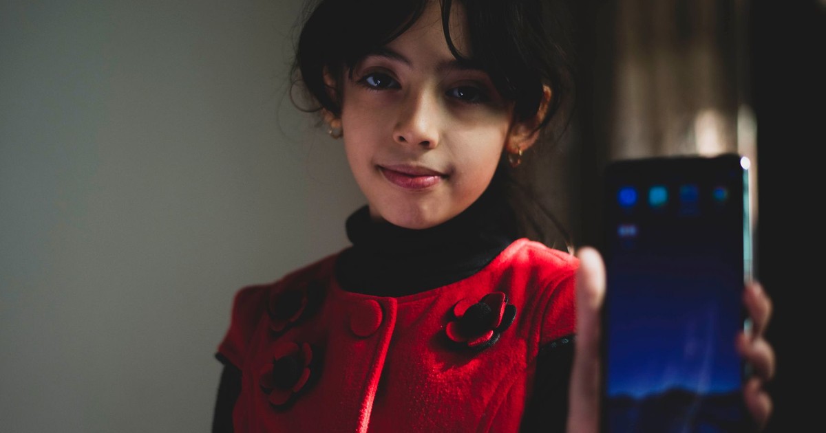 A young girl in a red dress shows a smartphone indoors, smiling softly.