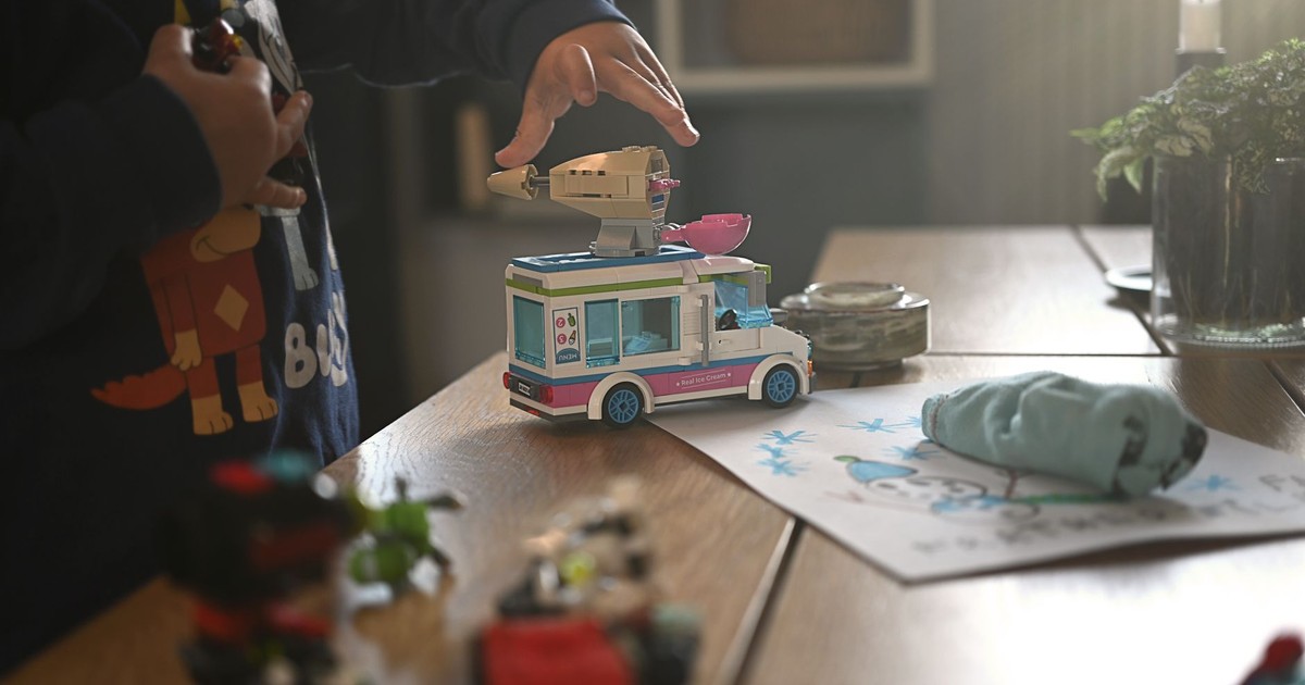 A child plays with a toy van on a well-lit table indoors, surrounded by colored drawings and action figures.