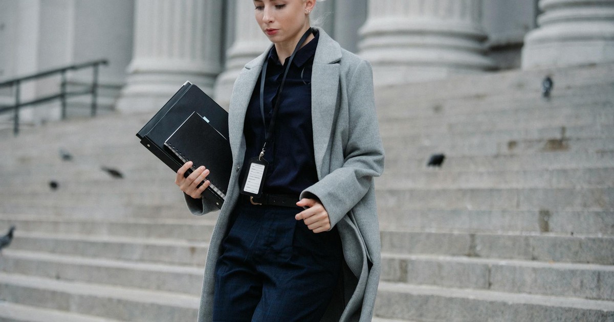 Young stylish female lawyer in formal clothes with name tag holding folders while going downstairs near stone building with columns