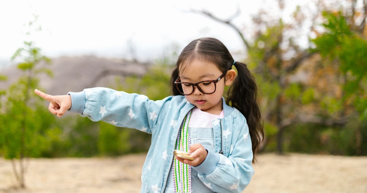 A curious young girl with glasses explores nature, holding a compass and pointing the way.