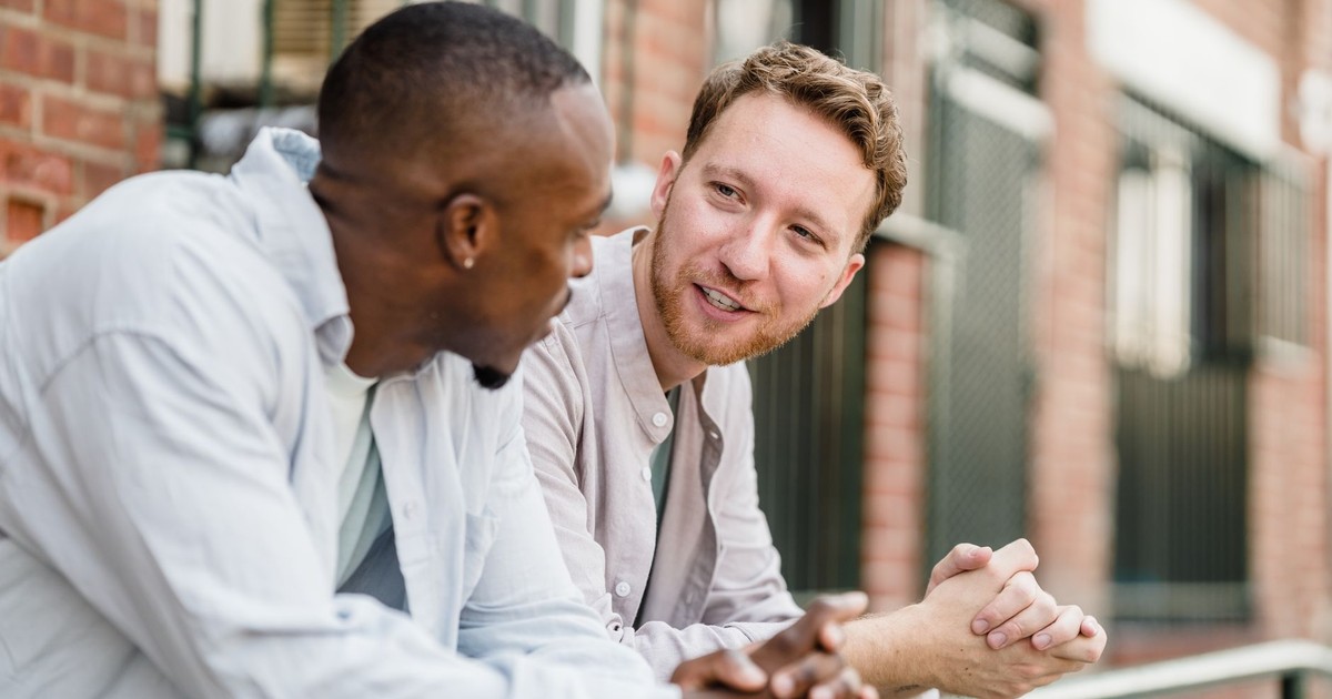 Two men engaging in lively conversation outdoors against a brick wall background.