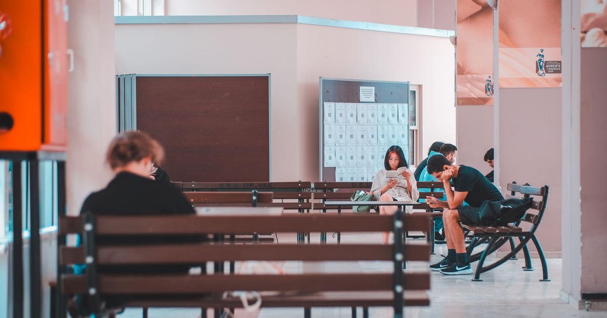 People sitting on benches in a school lobby, waiting and using smartphones, in a quiet indoor setting.
