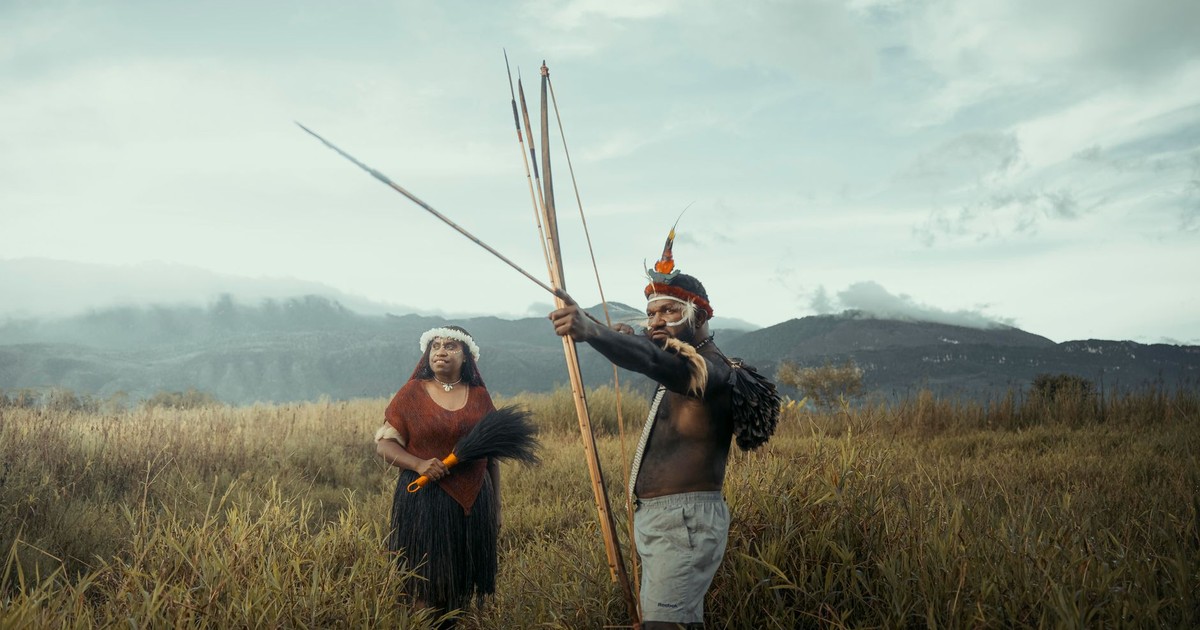 Indigenous duo holding traditional spears in a vast, green landscape with mountains.
