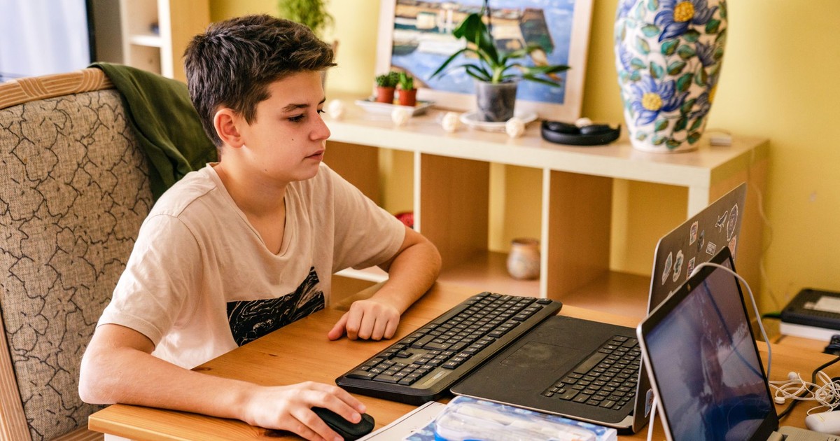 Teenage boy focused on computer work at a home desk setup with dual laptops and keyboard.