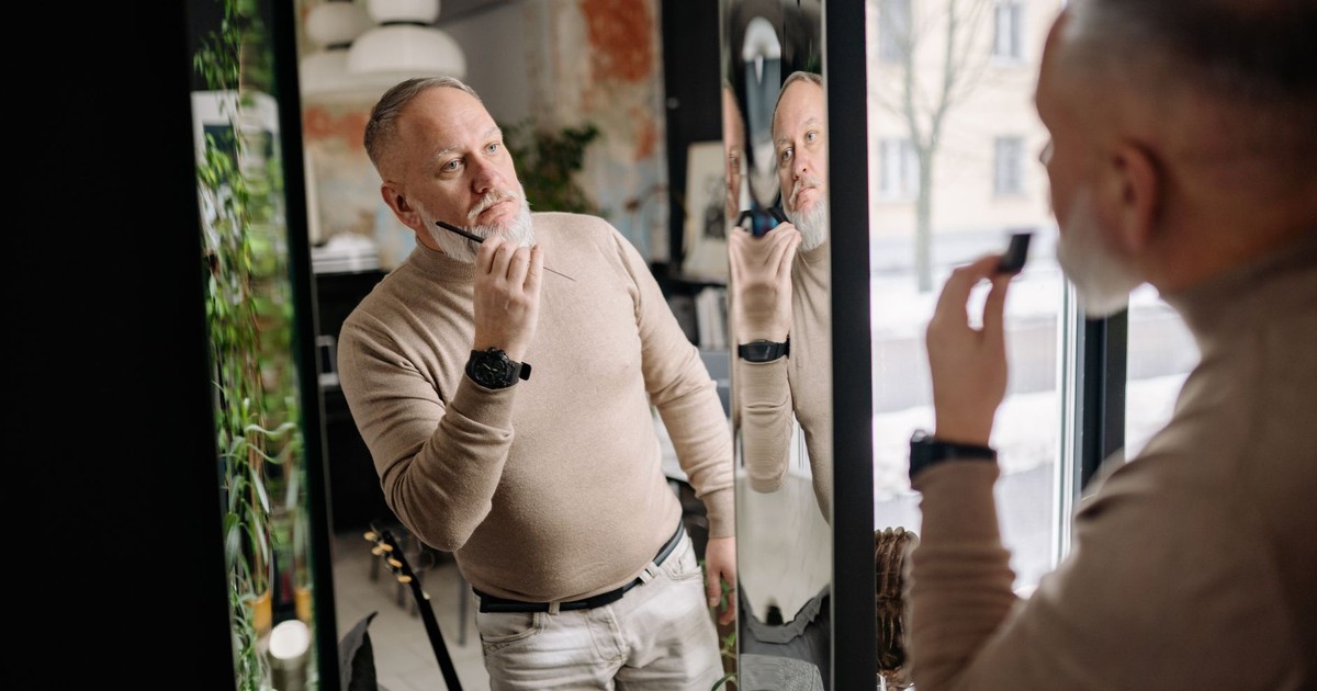 Middle-aged man applying beard care, examining reflection indoors, embracing modern grooming.