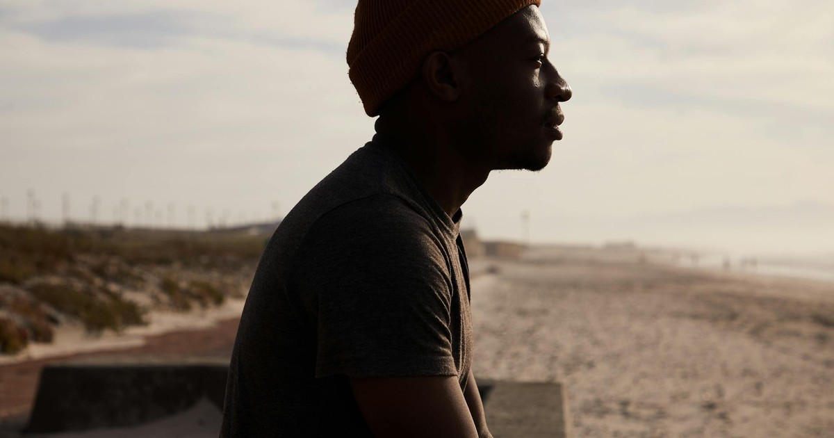 Side view of African American male in hat sitting on cement border on sandy shore near sea against cloudy sky