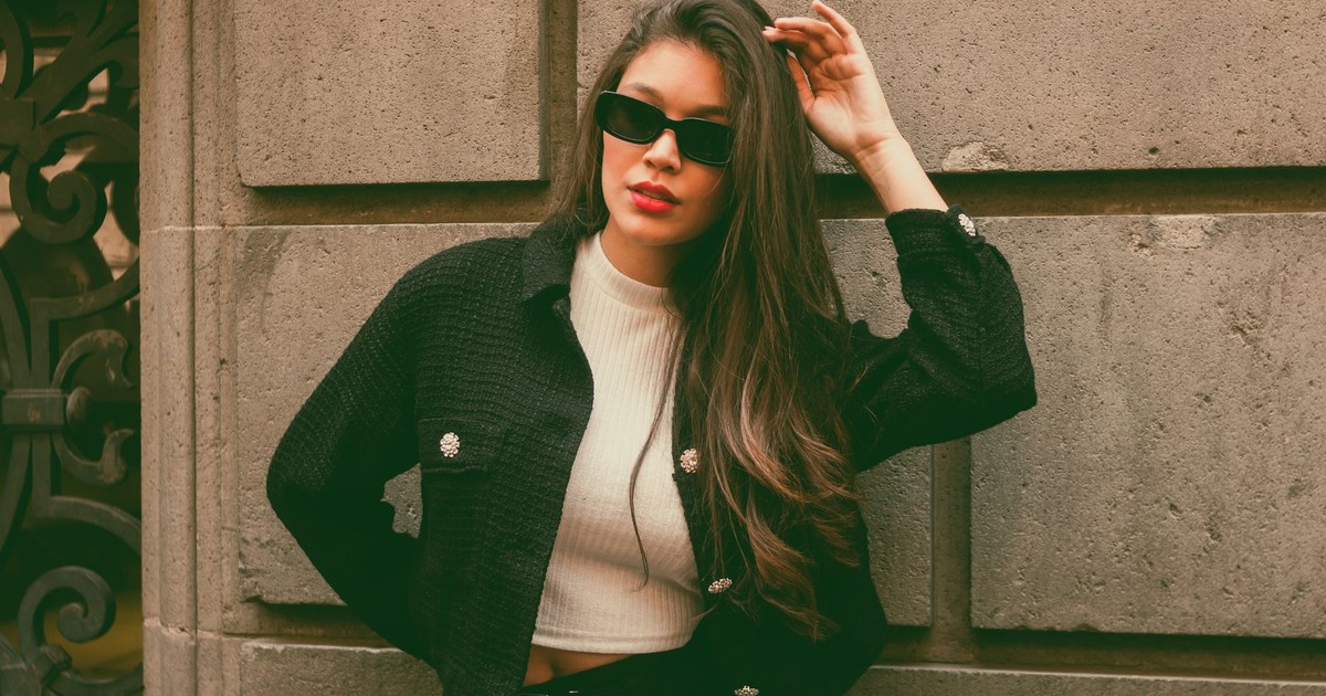 Trendy young woman wearing sunglasses and a black jacket poses confidently against a stone wall.