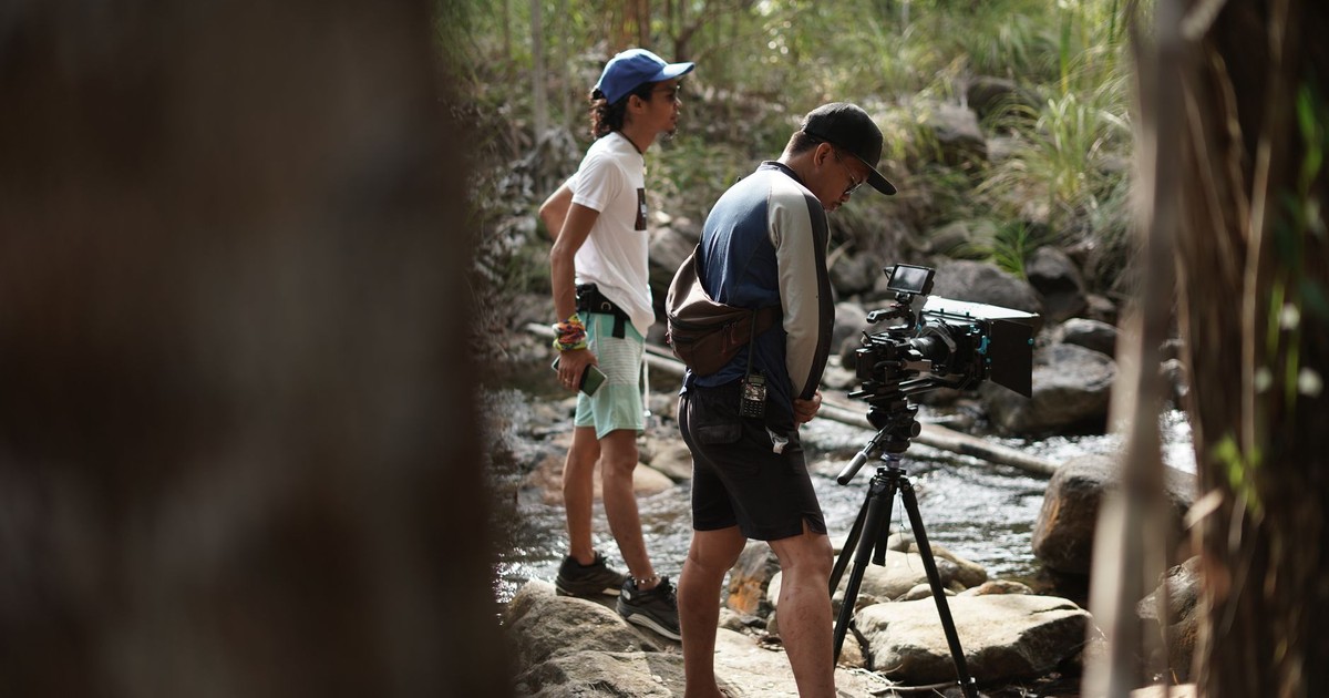 Two filmmakers prepare their camera equipment by a forest stream for a scene in natural surroundings.