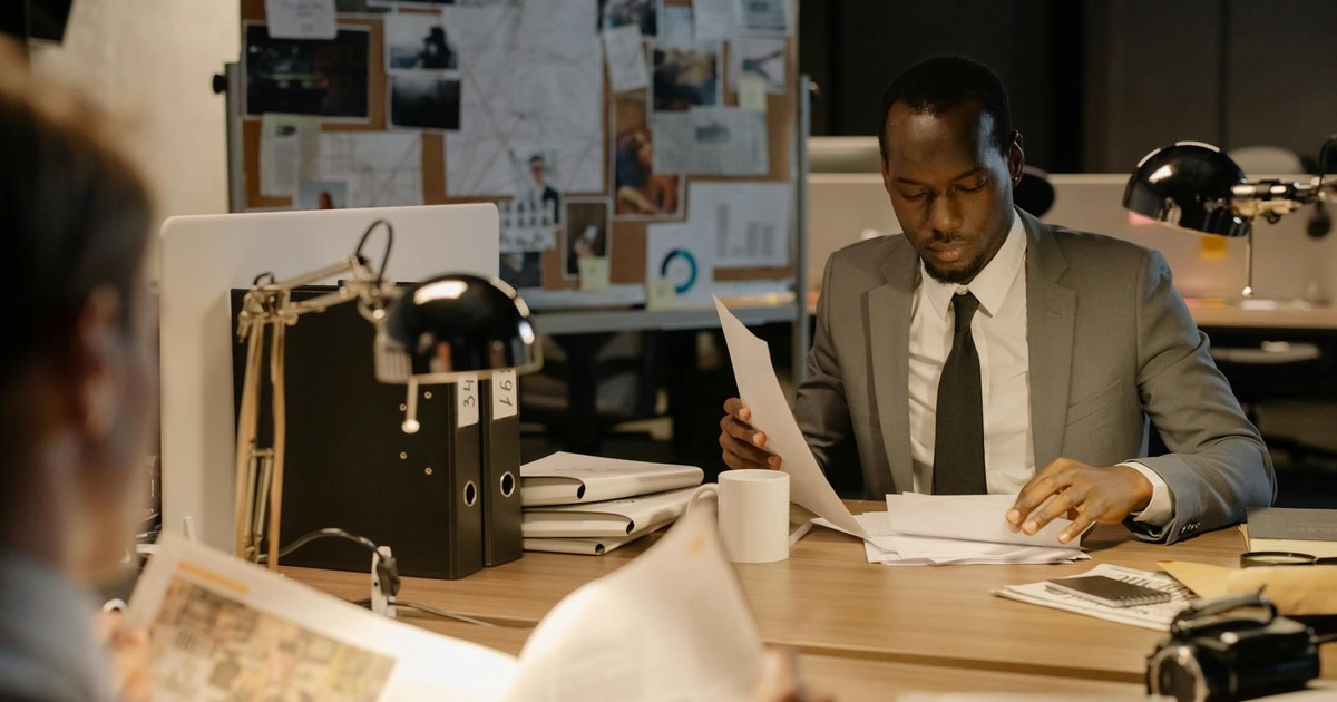 Detective in a suit examines papers at desk in a dimly-lit office, suggesting investigation work.