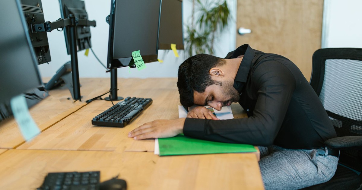 An exhausted office worker resting on a desk surrounded by computer monitors in a modern workspace.