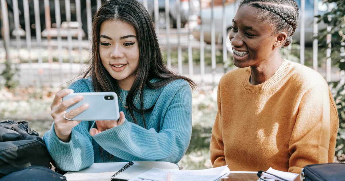 Happy young multiracial friends smiling and using mobile phone at table with notebooks on street