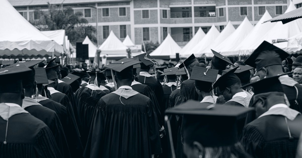 Diverse group of graduates in gowns at an outdoor ceremony with tents and buildings.
