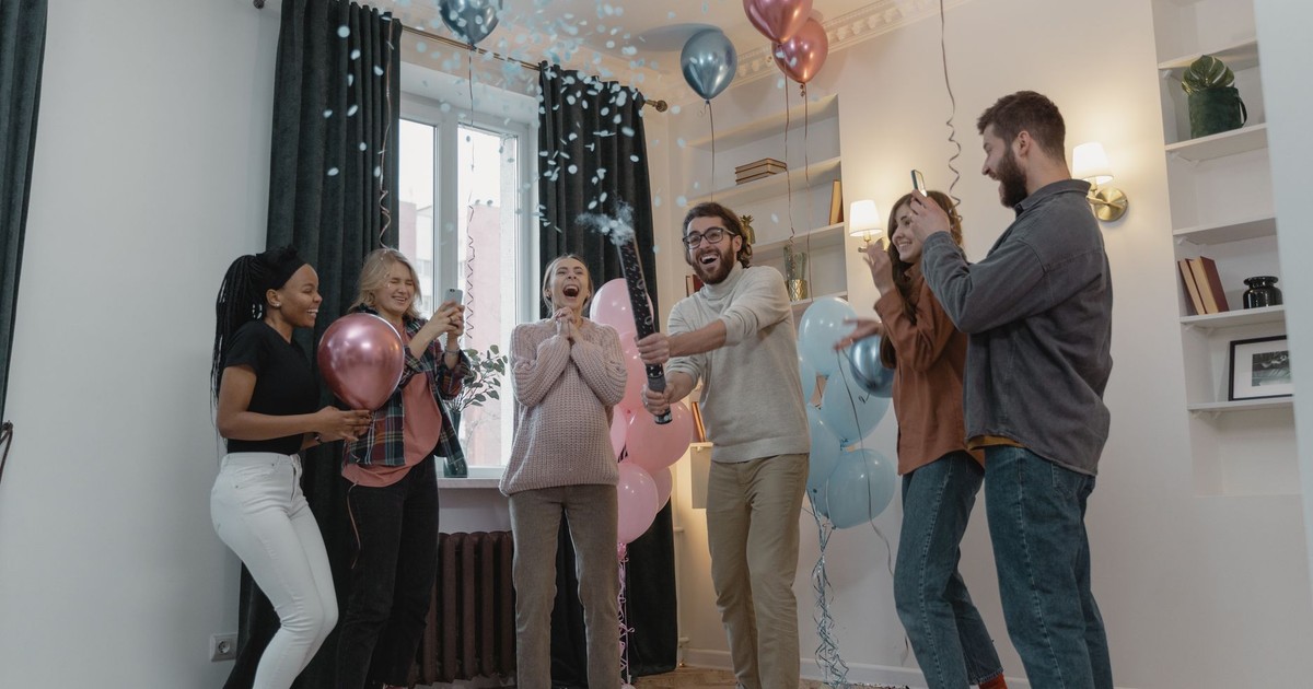 Excited friends celebrate a gender reveal with balloons and confetti indoors.