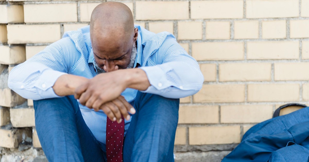 Sad shaved African American man in t shirt sitting with joined hands on background of brick wall and crying