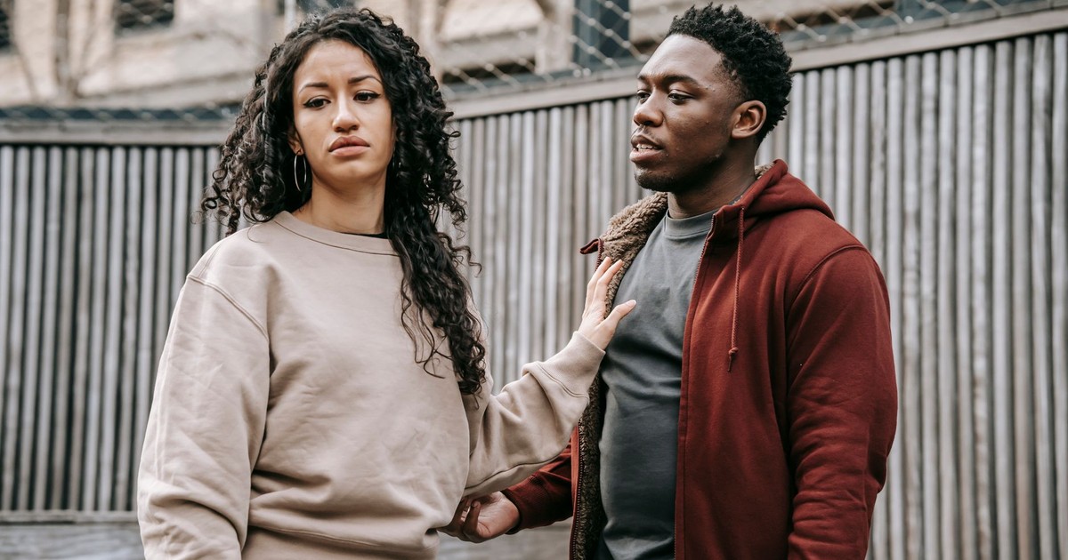 Sad ethnic girlfriend with curly hair rejecting annoyed African American boyfriend while arguing on street near wooden fence during breakup