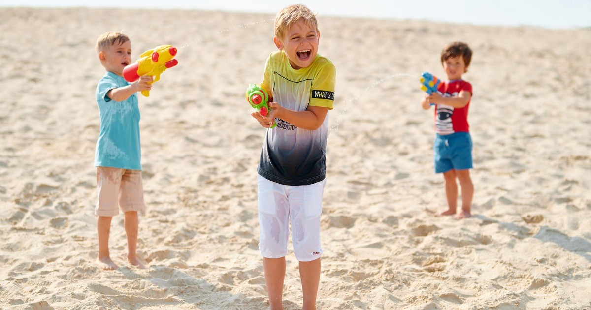 Three children joyfully playing with water guns on a sandy beach under the sun.
