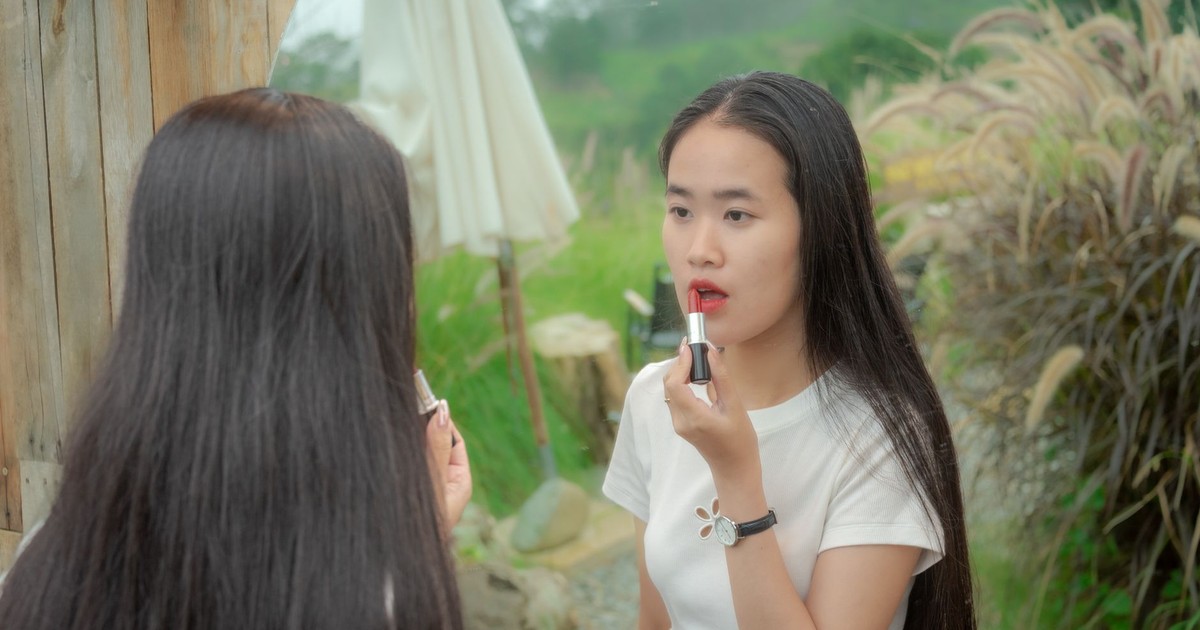 A young woman applies red lipstick using a mirror in an outdoor setting, creating a natural fashion moment.