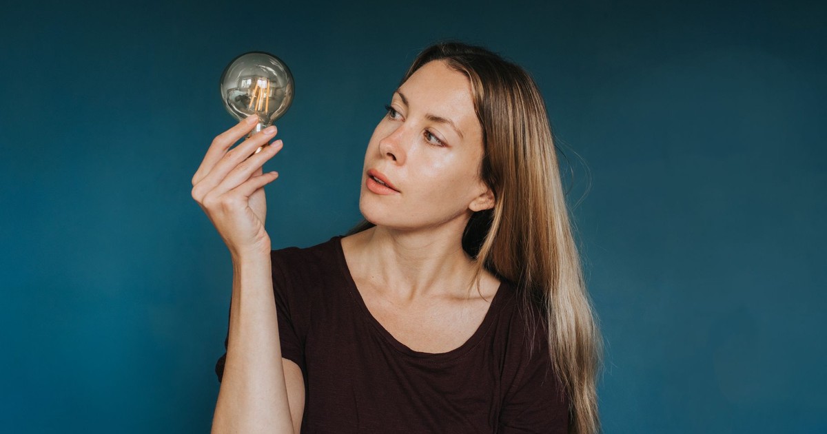 Enthusiastic female in casual clothes standing against blue background and looking at glowing light bulb