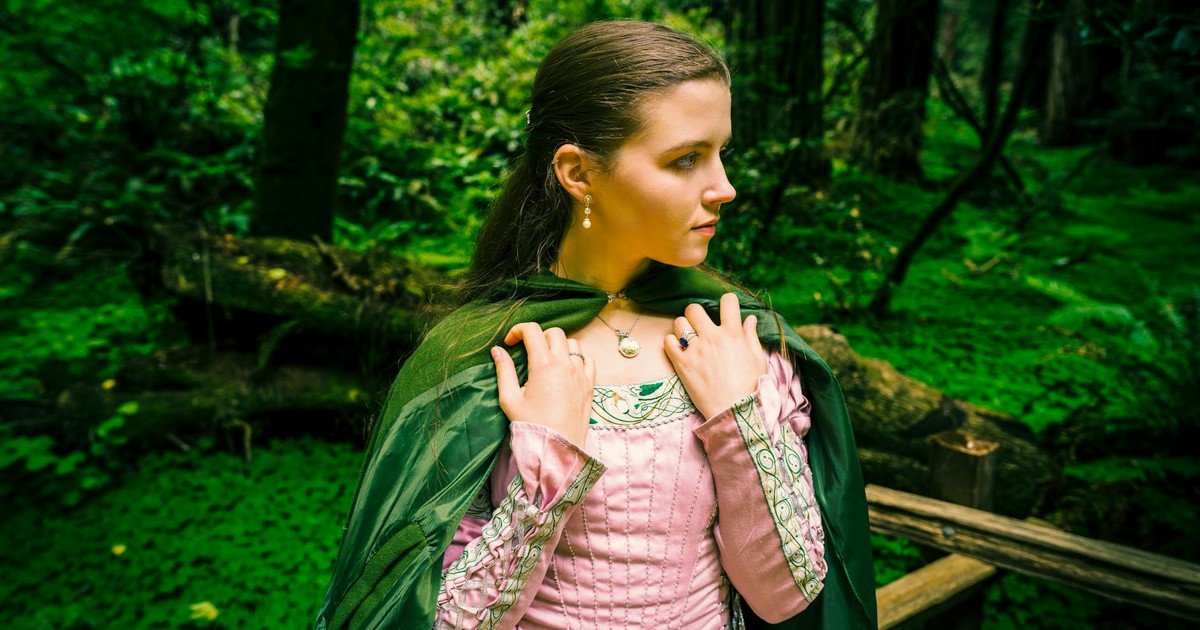 A woman in a pink dress and cloak stands in a lush forest, embodying a princess figure.