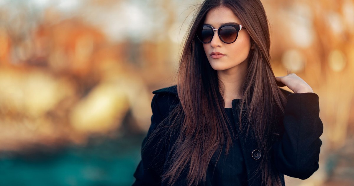 Fashionable woman wearing sunglasses and black coat, posing outdoors in natural light.