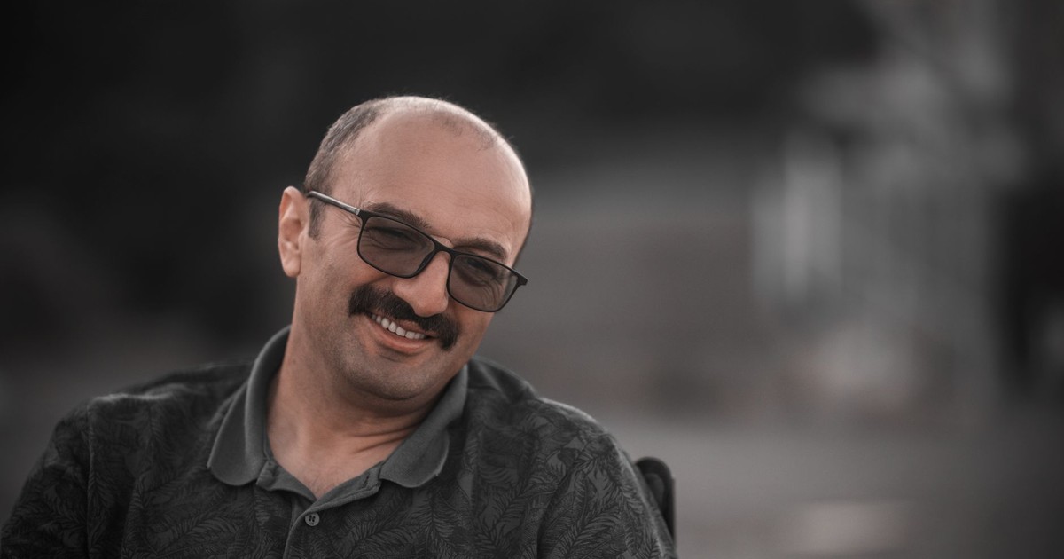 Man with eyeglasses smiles warmly outdoors, wearing a gray polo shirt.