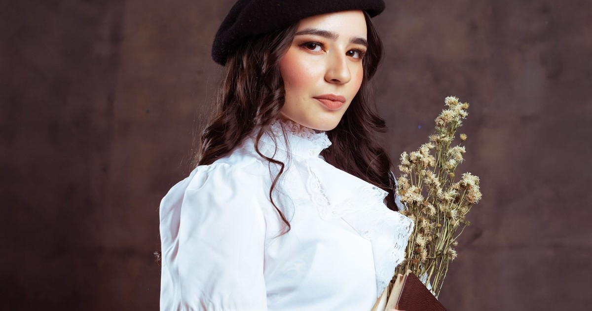 Portrait of an elegant woman in a white blouse and beret, holding flowers against a studio backdrop.