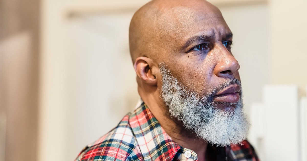 A thoughtful portrait of a bald, bearded man in a plaid shirt, looking away indoors.