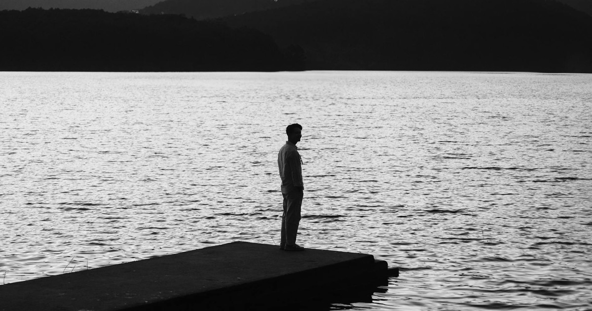 A lone man stands on a dock, gazing across a tranquil lake against a backdrop of mountains.