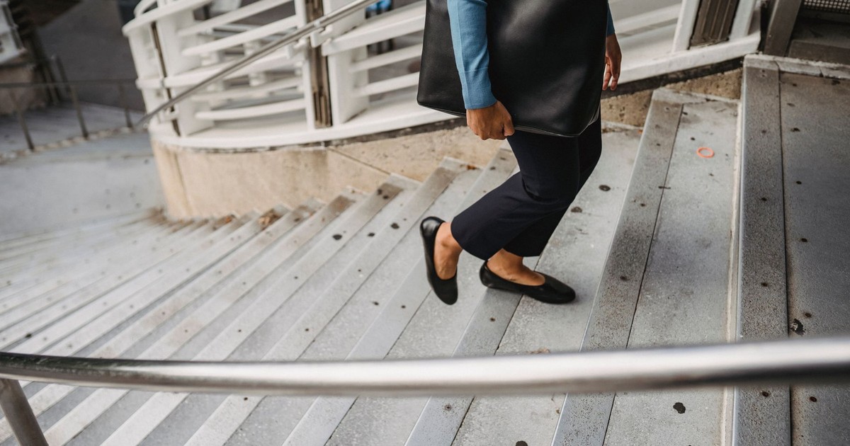 Unrecognizable female in casual clothes and black leather bag walking up concrete staircase with metal railing on street in city