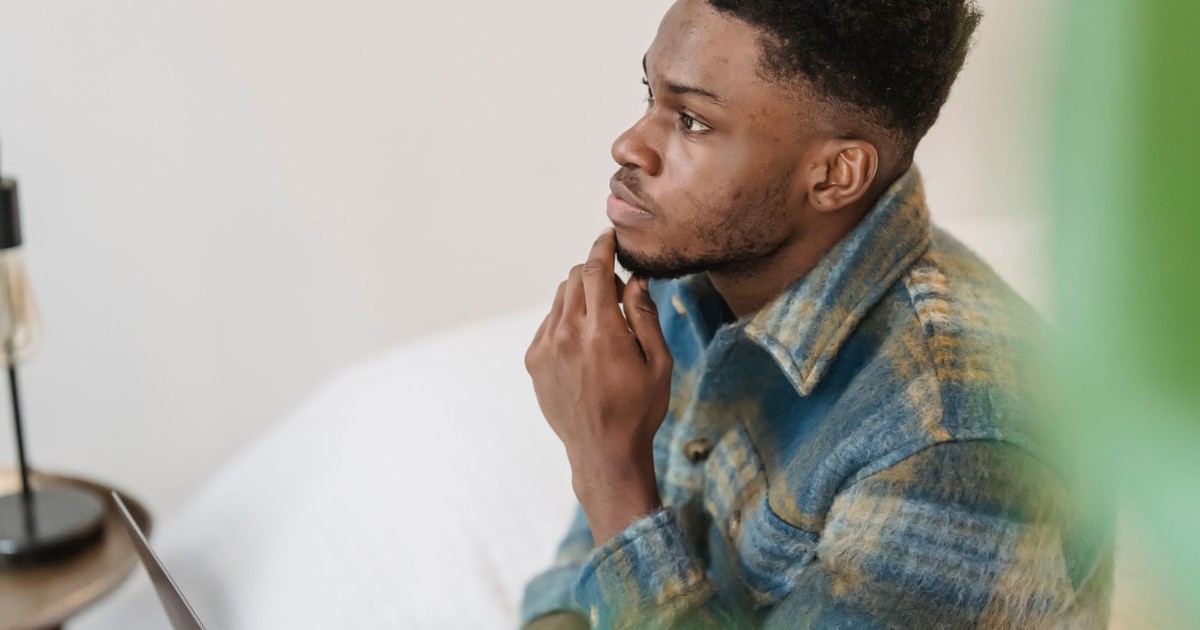 Side view of young African American male in stylish shirt looking away pensively and touching chin while sitting at home and thinking