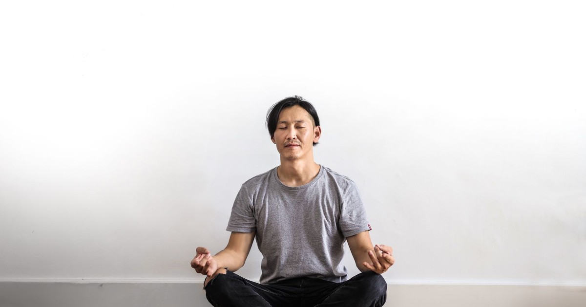 Asian man practicing meditation indoors on a wooden floor, promoting relaxation and mindfulness.