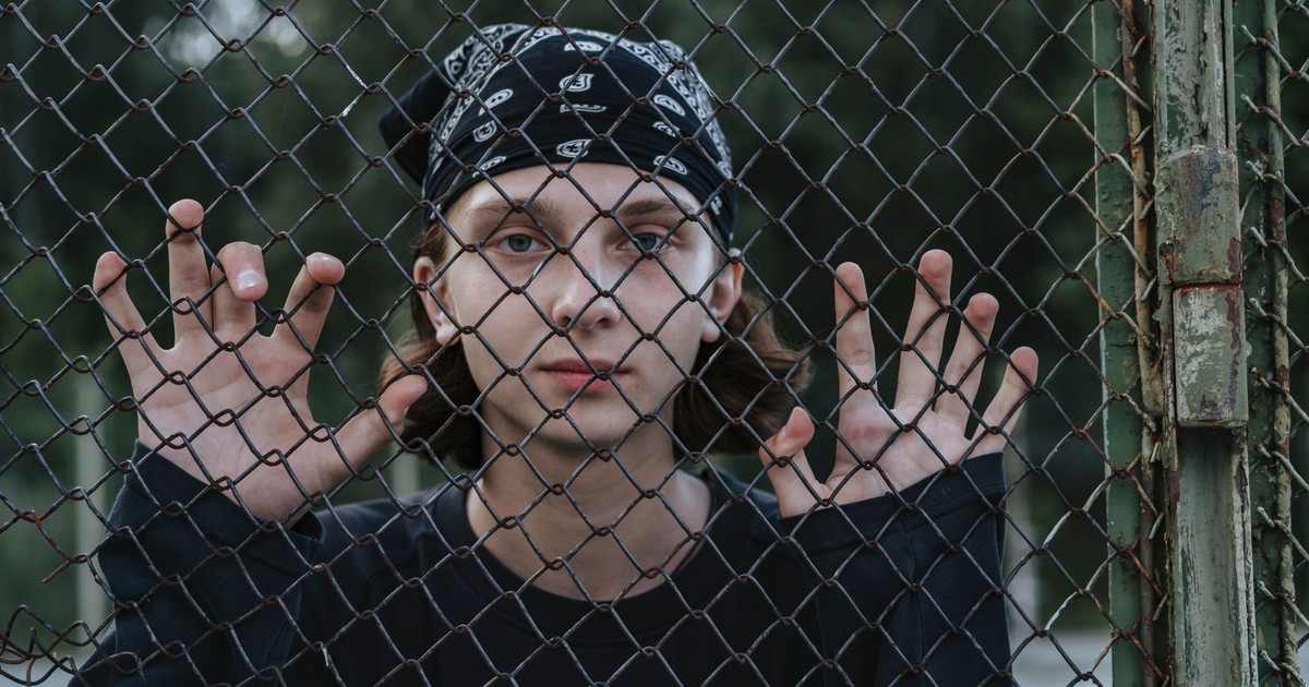Teen wearing black bandana and long sleeves poses behind a chain link fence, symbolizing youth expression.
