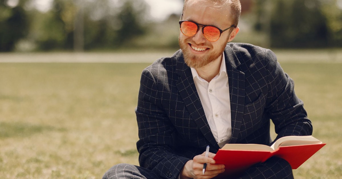 A cheerful man in sunglasses enjoys reading a book in the park on a sunny day, exuding happiness and relaxation.