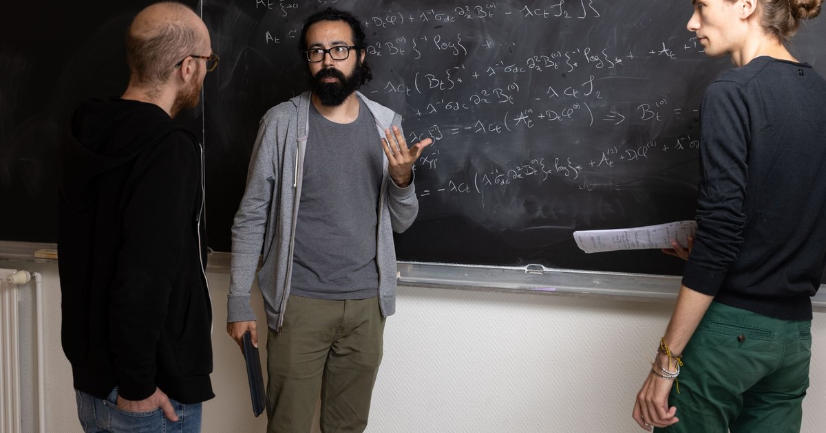Three adults solving complex mathematical equations on a chalkboard indoors.