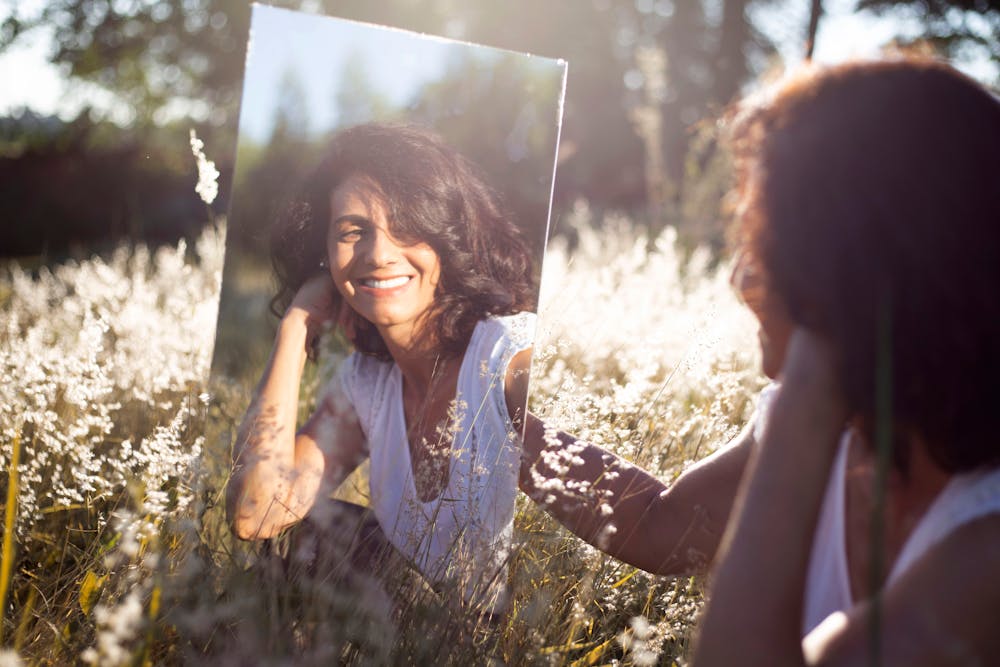 A woman smiles at her reflection in a mirror placed in a sunny field