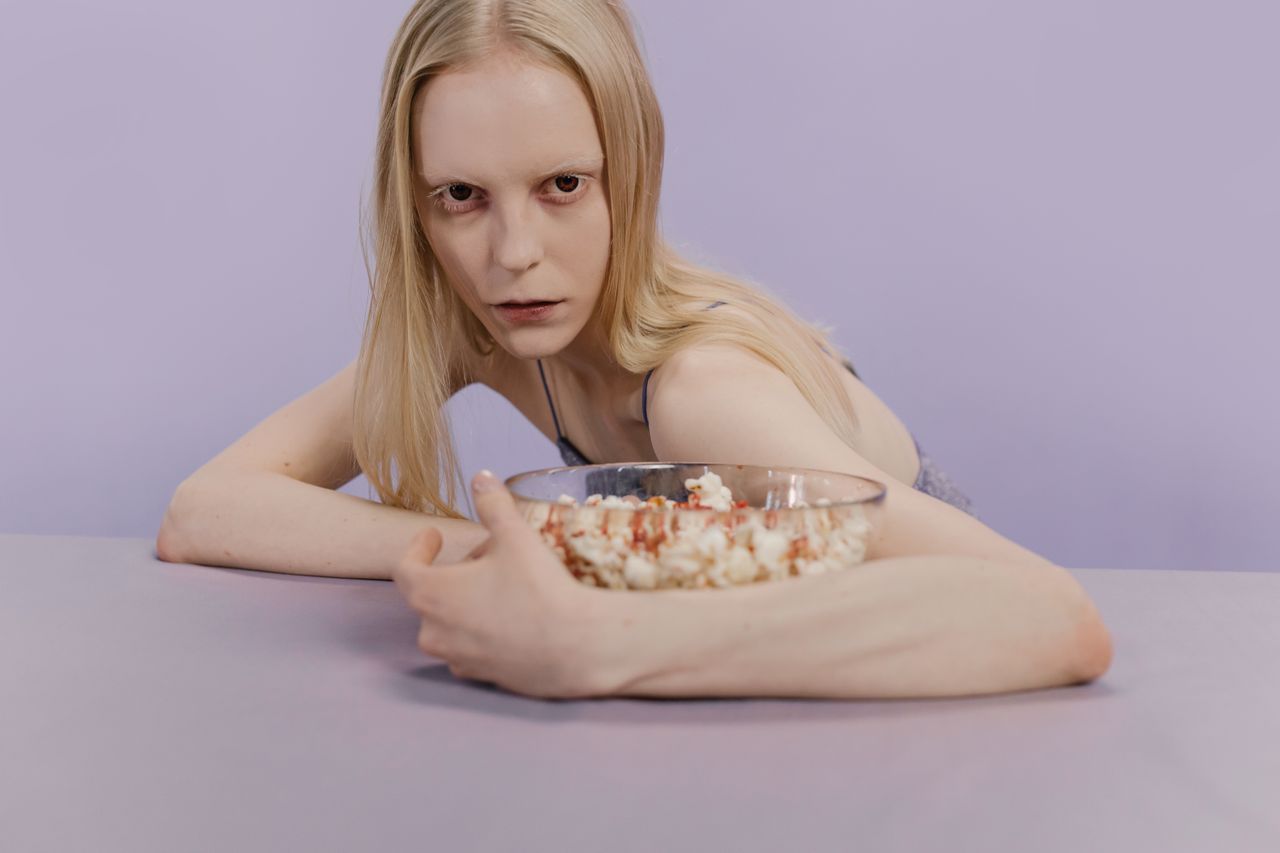 A woman with a neutral expression holds a bowl of popcorn on a minimalist pastel backdrop.