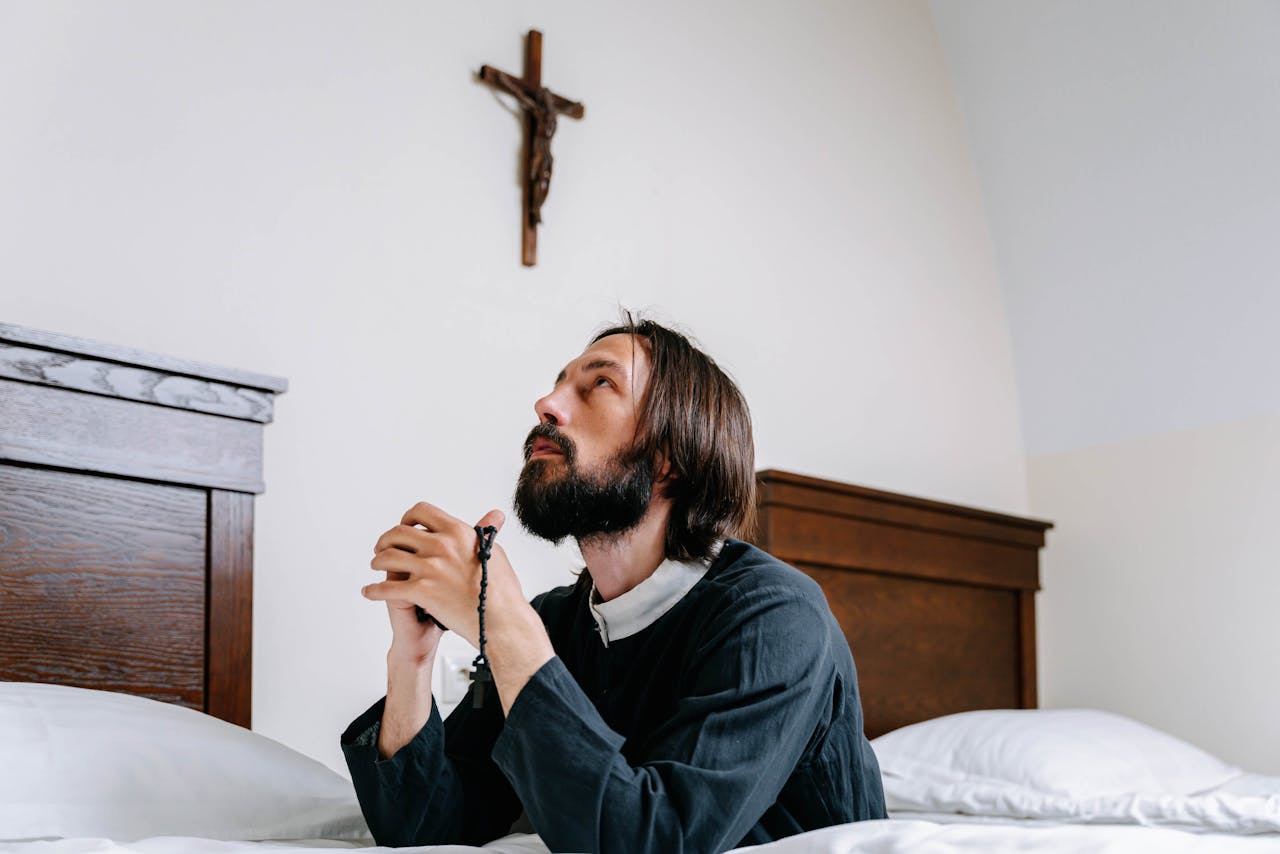 A man kneeling in prayer with a rosary before a crucifix in a cozy bedroom setting