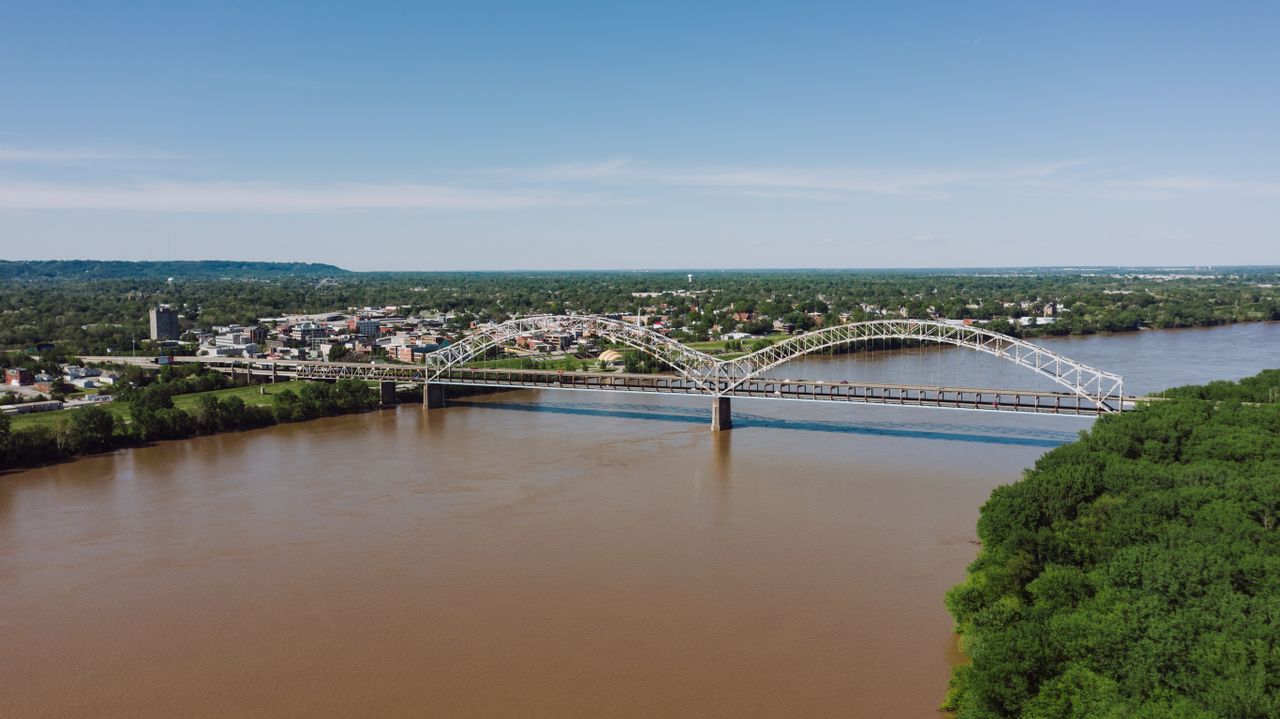 A scenic aerial view of a bridge crossing a river in Louisville, KY, with lush greenery and a clear blue sky