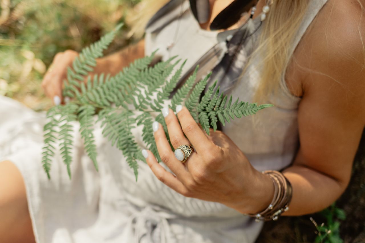 Close-up of a woman holding a fern leaf, wearing a white dress and jewelry outdoors