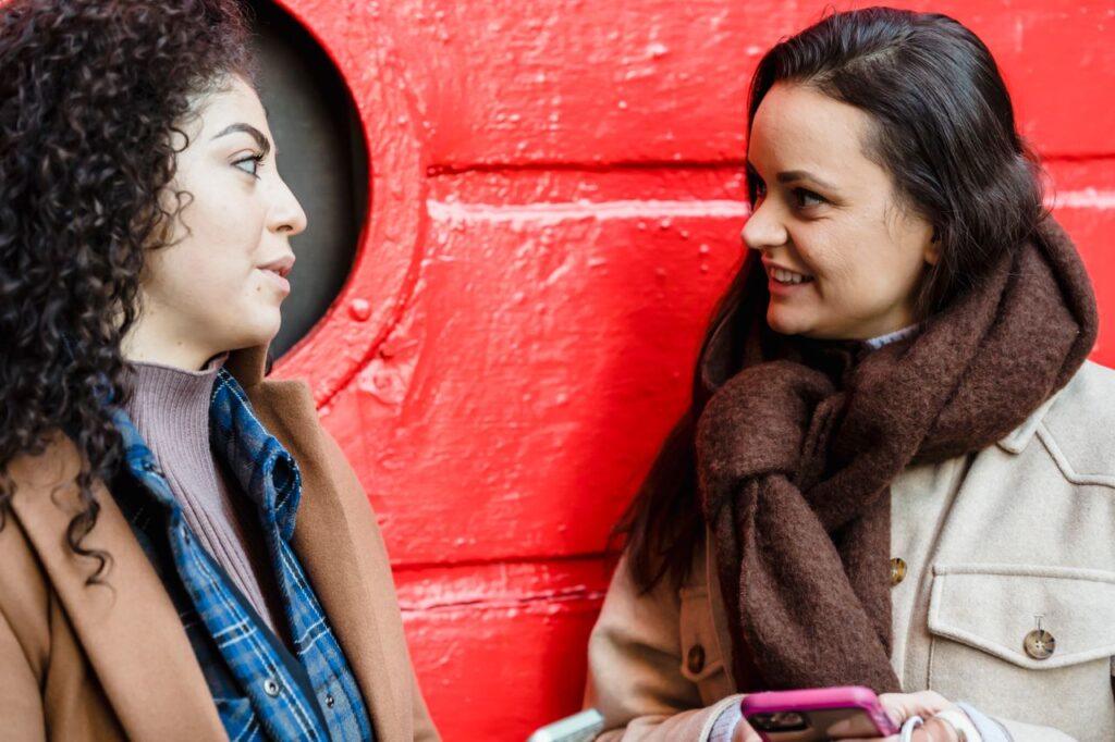 Two young women having a friendly conversation outdoors against a vibrant red backdrop