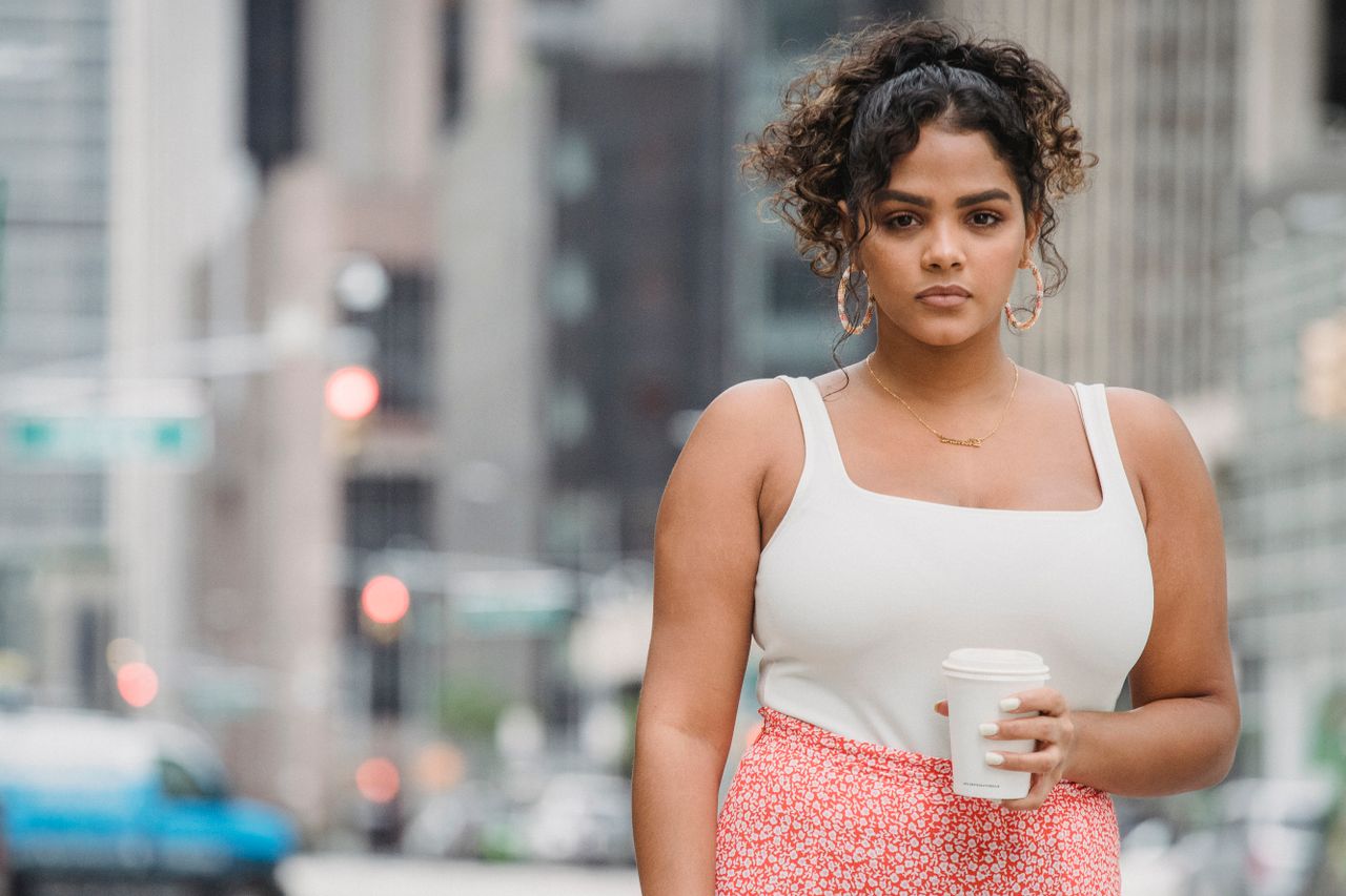 A confident woman stands outdoors in a city holding a coffee cup, exuding style and individuality.