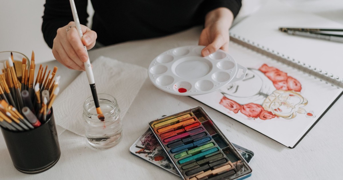 Unrecognizable painter with palette and paintbrush sitting at table with sketchbook and art supplies while painting in modern light studio