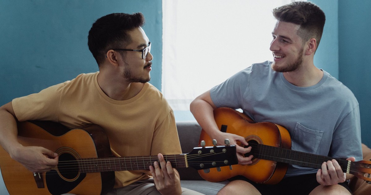 Two men happily playing acoustic guitars together in a bright living room setting.